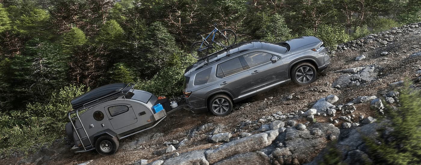 A dark grey 2024 Honda Pilot is shown towing a small camper trailer on a steep rocky hill.