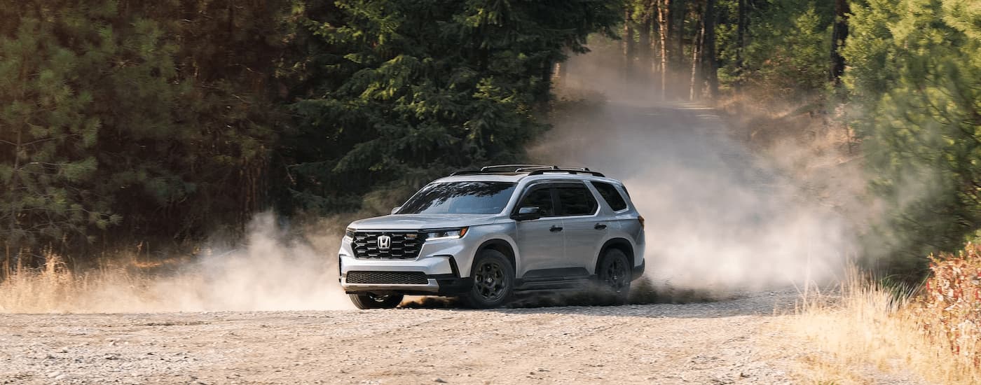 A silver 2024 Honda Pilot Trailsport is shown driving on a dusty dirt road.