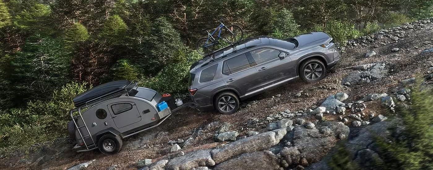 A grey 2023 Honda Pilot Touring is shown towing a small camper trailer on a rocky road.